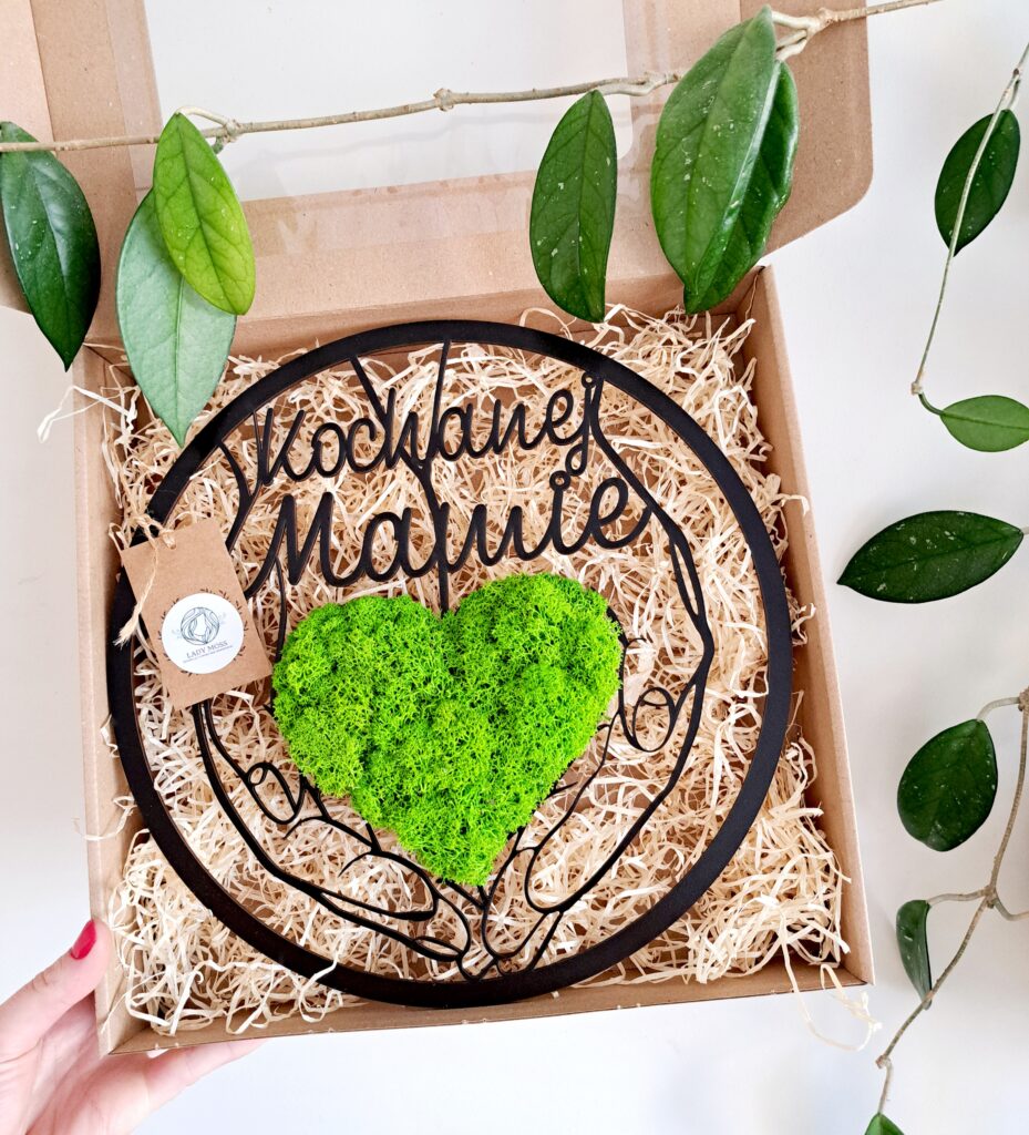 Dekoracja z chrobotka reniferowego w kształcie serca trzymanego w dłoniach, umieszczona w obręczy 30 cm, z napisem „Kochanej Mamie”.